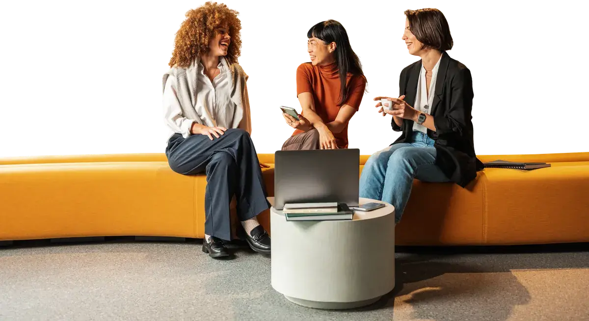 Three women talk on a lounge bench with coffee and a laptop